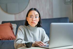 Korean woman with perplexed face, sitting with laptop, working on remote, freelancer with confused expression looking at camera photo