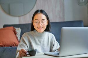 Close up portrait of beautiful asian woman drinks her coffee, smells drink in cup, takes brake from working or studying on laptop photo