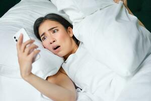 Portrait of asian woman waking up in bed, looking shocked at mobile phone, realise she overslept photo