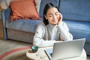 hermosa asiático niña trabajos desde hogar, estudiando con computadora portátil, sentado en vivo habitación, siendo en remoto, independiente en línea foto