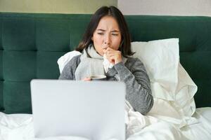 Portrait of sick asian woman watching videos on laptop, staying in bed and coughing, catching cold photo