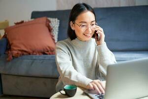 Portrait of smiling asian woman making phone call, working on laptop and having conversation on smartphone photo