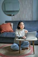 Vertical shot of korean working woman, sitting on floor at home with laptop, studying, using computer on remote, e-learning concept photo