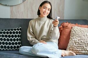 Beautiful asian girl in cozy home, watching television, holding remote and switching channels, sitting on sofa photo