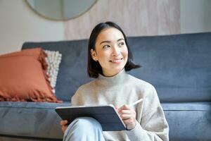 Portrait of asian woman with tablet, drawing, working on design project, holding pen, sitting in her living room, freelancing photo