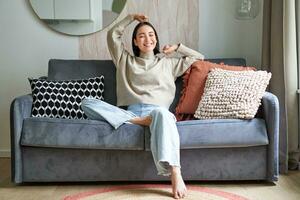 Portrait of happy asian woman feeling lazy, stretching on sofa and smiling pleased, relaxing at home, resting from work photo