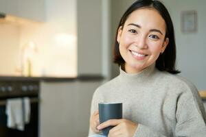 retrato de sonriente asiático niña con taza de café, Bebiendo caliente té y consiguiendo acogedor a hogar. calentamiento arriba con bebida foto