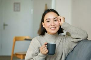 Smiling asian woman sitting at home with cup of coffee, relaxing and feeling warmth, looking outside window, resting on sofa in living room photo