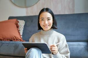 Portrait of asian woman with tablet, drawing, working on design project, holding pen, sitting in her living room, freelancing photo