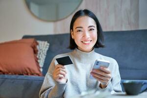 Portrait of smiling korean woman using credit card and smartphone app, paying for purchase, order online delivery photo