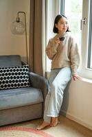 Vertical shot of beautiful woman sitting in her house, looking outside window and drinking coffee photo