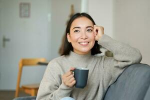 Smiling asian woman sitting at home with cup of coffee, relaxing and feeling warmth, looking outside window, resting on sofa in living room photo