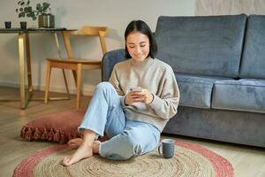 Image of stylish young woman in modern house, using mobile phone, sitting on floor and holding smartphone, drinking from cup photo