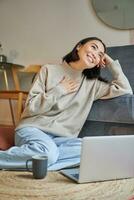 Portrait of dreamy woman sitting with laptop on floor, watching on computer and drinking coffee, enjoying cozy days at home photo