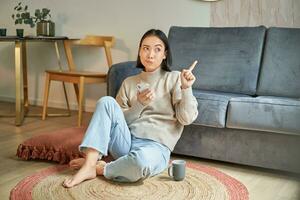 Portrait of woman sitting on floor with smartphone, looking thoughtful and pointing finger at banner, promo advertisement on top right corner photo