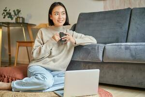 Image of smiling asian woman drinking hot tea, holding cup and sitting near laptop on floor, resting at home photo