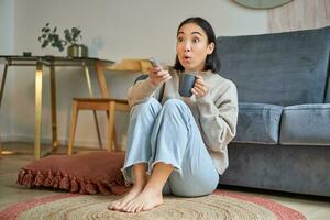 Portrait of girl watching television at home, sits on floor near sofa, holds remote and changes channels photo