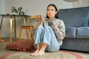 Image of young asian woman with remote, watching tv, drinking coffee, looking suspicious at television screen photo