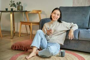 retrato de elegante coreano mujer se sienta en piso con teléfono inteligente, utilizando móvil teléfono, sonriente complacido, concepto de quedarse a hogar y relajación foto