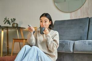 Portrait of young korean woman watching television, holding remote and looking amazed at tv screen, spending time at home photo