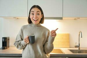 Portrait of smiling asian girl standing in kitchen, drinking coffee from cup and pointing at banner, showing letting agencies advertisement photo