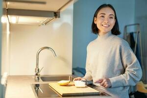 Cute asian woman making herself toast, cut loaf of bread, preparing sandwitch on kitchen photo