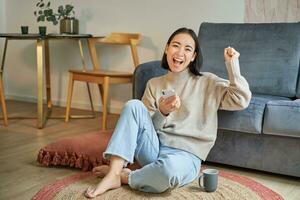 Enthusiastic asian woman sits on floor with smartphone, raise hand up and cheering, celebrating victory photo