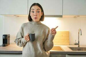 Portrait of smiling asian girl standing in kitchen, drinking coffee from cup and pointing at banner, showing letting agencies advertisement photo