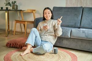 Smiling asian girl sits on floor in stylish living room, pointing finger at advertisement, showing promo banner, holding mobile phone in hand photo