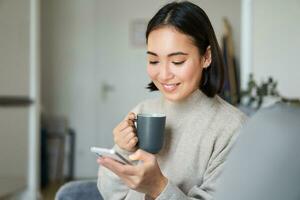 retrato de sonriente asiático niña comprobación su Noticias alimentar en teléfono inteligente y Bebiendo café, sentado en sofá a hogar, hojeada en móvil teléfono, leyendo foto