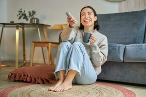 Image of young korean woman drinks coffee, holds remote from tv, watching television at home, resting in her house photo