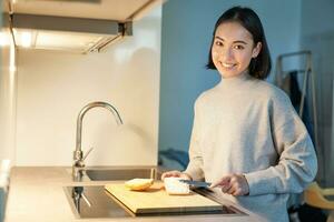 Smiling asian woman cooking sandwitch, cut loaf of bread in kitchen and looking at camera photo