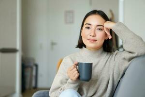 Smiling asian woman sitting on sofa with her mug, drinking coffee at home and relaxing after work, looking calm and cozy photo