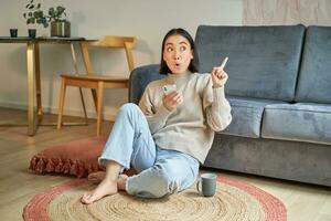 Portrait of modern young korean woman sitting on floor, pointing finger at copy space, showing banner or advertisement, holding smartphone as using app photo