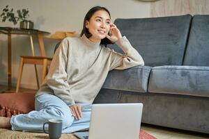 Image of smiling asian woman drinking hot tea, holding cup and sitting near laptop on floor, resting at home photo