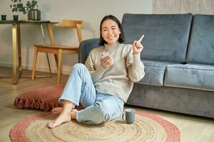 Portrait of modern young korean woman sitting on floor, pointing finger at copy space, showing banner or advertisement, holding smartphone as using app photo
