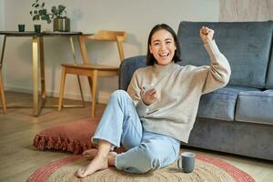 Enthusiastic asian woman sits on floor with smartphone, raise hand up and cheering, celebrating victory photo