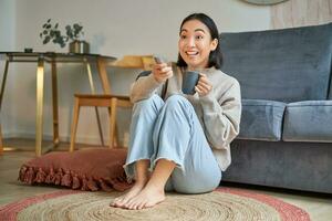 Portrait of girl watching television at home, sits on floor near sofa, holds remote and changes channels photo