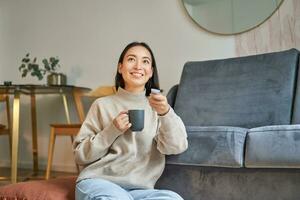 retrato de sonriente coreano mujer sentado cerca televisor, participación remoto y traspuesta canales mientras Bebiendo caliente café foto