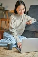 Vertical shot of young woman in cozy home working on laptop, using smartphone and drinking coffee, sitting on floor near sofa photo