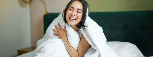 Beautiful asian woman sitting on bed, covered with white duvet, smiling, enjoying happy weekend morning, laughing at camera photo
