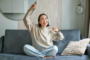 Happy and carefree girl singing and listening to music on smartphone app, using mobile phone as microphone, sitting on sofa photo