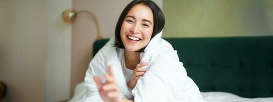 Happy beautiful asian woman covered with blanket, sits on bed in warm duvet, tries to reach camera with a hand, smiling playfully. Hotel bedroom, comfort and apartments concept photo