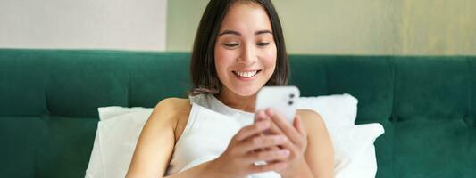 Portrait of smiling asian woman lying in bed, looking at smartphone, using mobile phone with happy face, relaxing on lazy morning photo