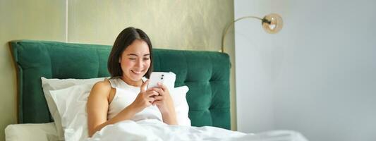 hermosa sonriente asiático mujer en cama, leyendo mensaje en teléfono inteligente y reír, disfrutando perezoso día en dormitorio, utilizando móvil teléfono foto