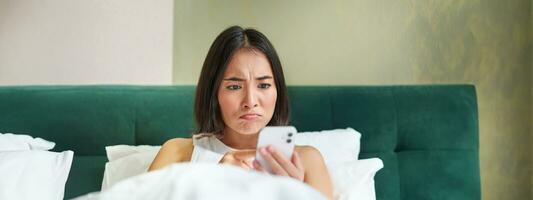 Close up portrait of grumpy asian girl lying in bed, grimacing and pouting from complicated issue on her mobile phone, reading smartphone app photo