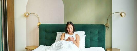 Portrait of woman lying in bed under duvet, looking at smartphone with serious face, reading message, watching news on mobile phone app photo