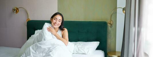 contento coreano mujer despierta arriba en su hotel habitación, acostado en cama debajo acogedor calentar mantas, blanco hojas, disfrutando fin de semana Mañana foto