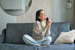 Portrait of carefree asian girl sits at home, listens to music in headphones and sings at smartphone, holds mobile phone like microphone photo