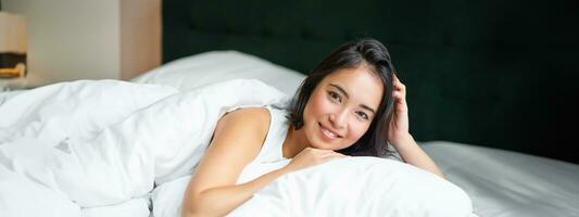 retrato de joven coreano mujer sonriente, acostado en cama en almohada, posando en su dormitorio, mirando relajado y sensación cómodo foto